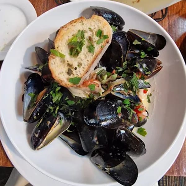 A white bowl of mussels with broth, garnished with parsley and a slice of toasted bread.