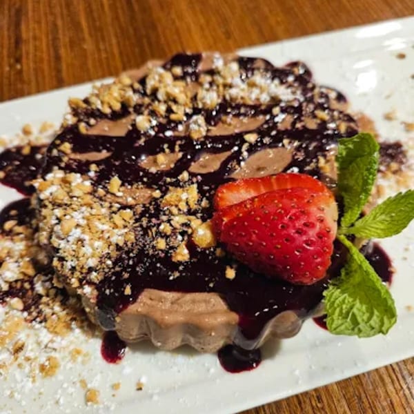Chocolate dessert with berry sauce, nuts, powdered sugar, strawberry, and mint on a white plate.